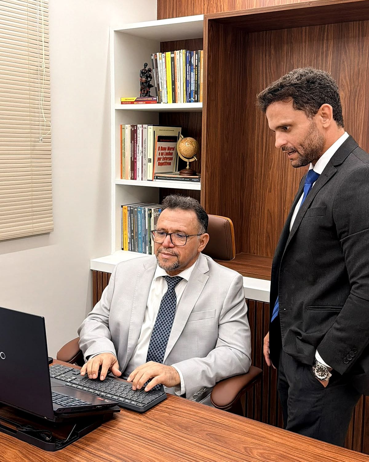 Vianey Martins, sentado ao computador, e Bruno Rafael Gomes Silva, de pé, trabalhando no escritório diante de uma estante de livros jurídicos.